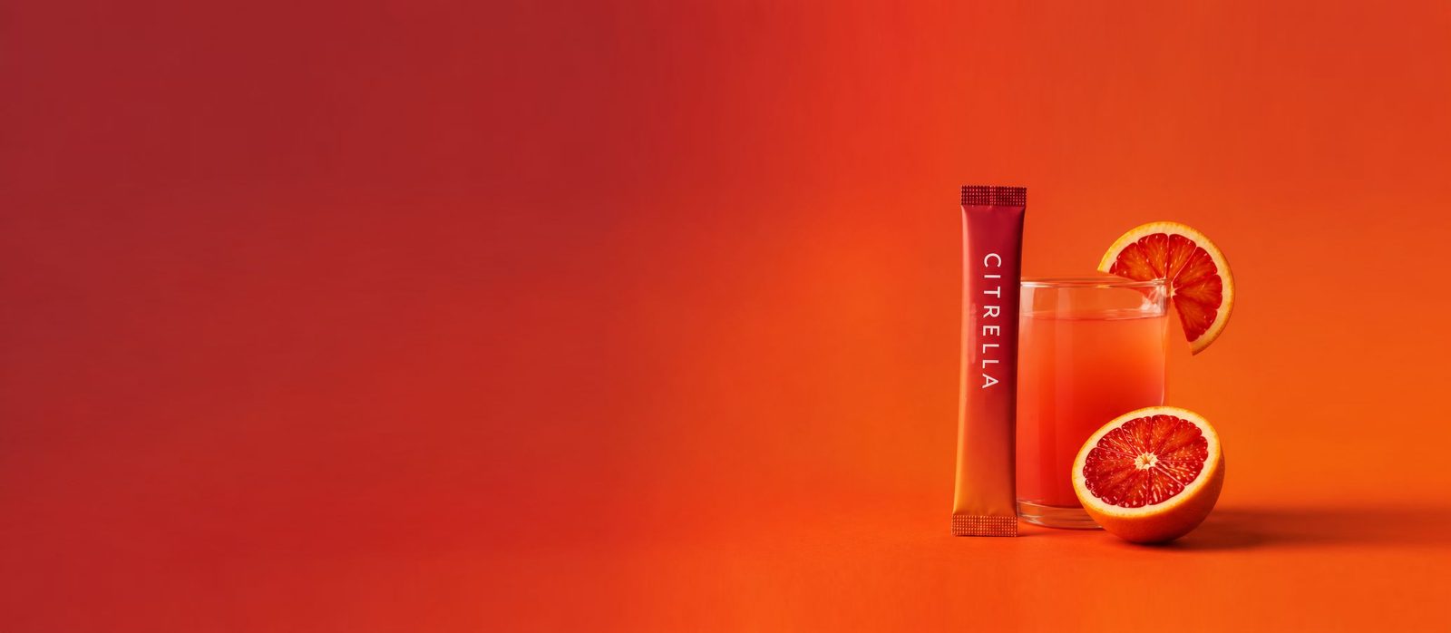 A Citrella slim stick standing beside a glass of blood orange drink with fresh orange halves